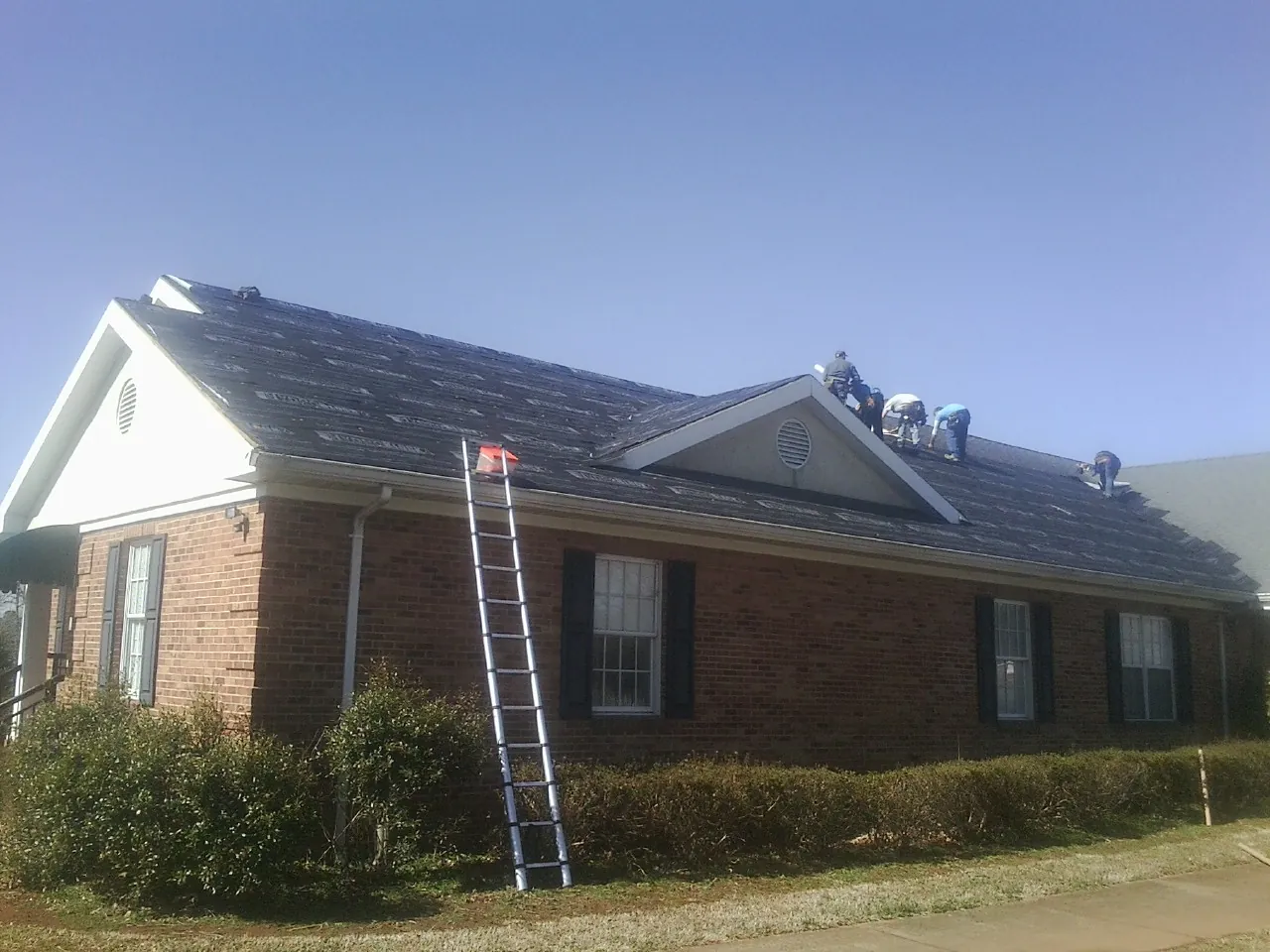 Crew tearing shingles off a brick ranch roof with a ladder set — Metal Roof Repair work by our crew in Barnardsville