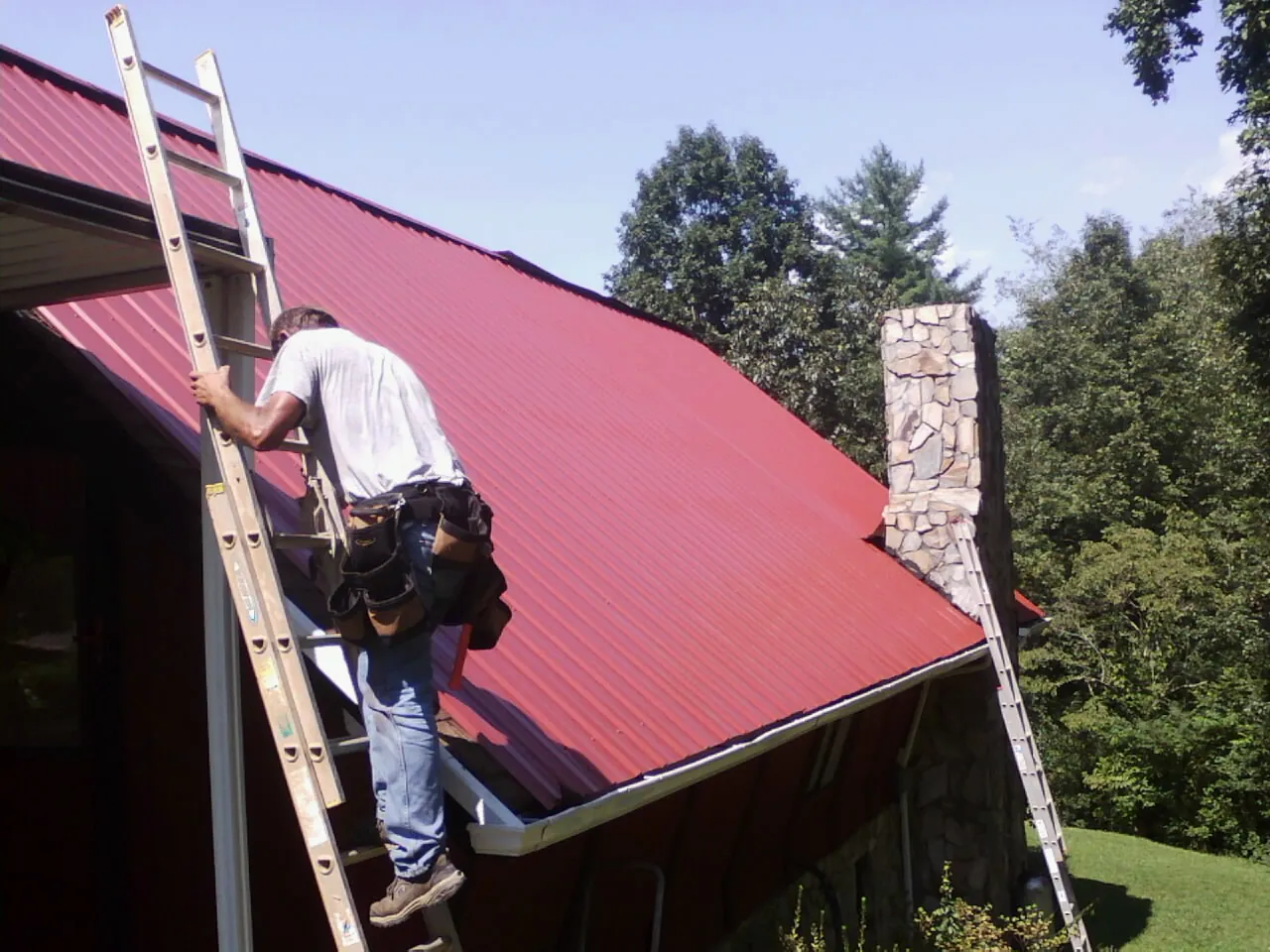 Roofer on a ladder beside a red metal roof and stone chimney — Soffit & Fascia Repair by Roofing Brothers in Barnardsville